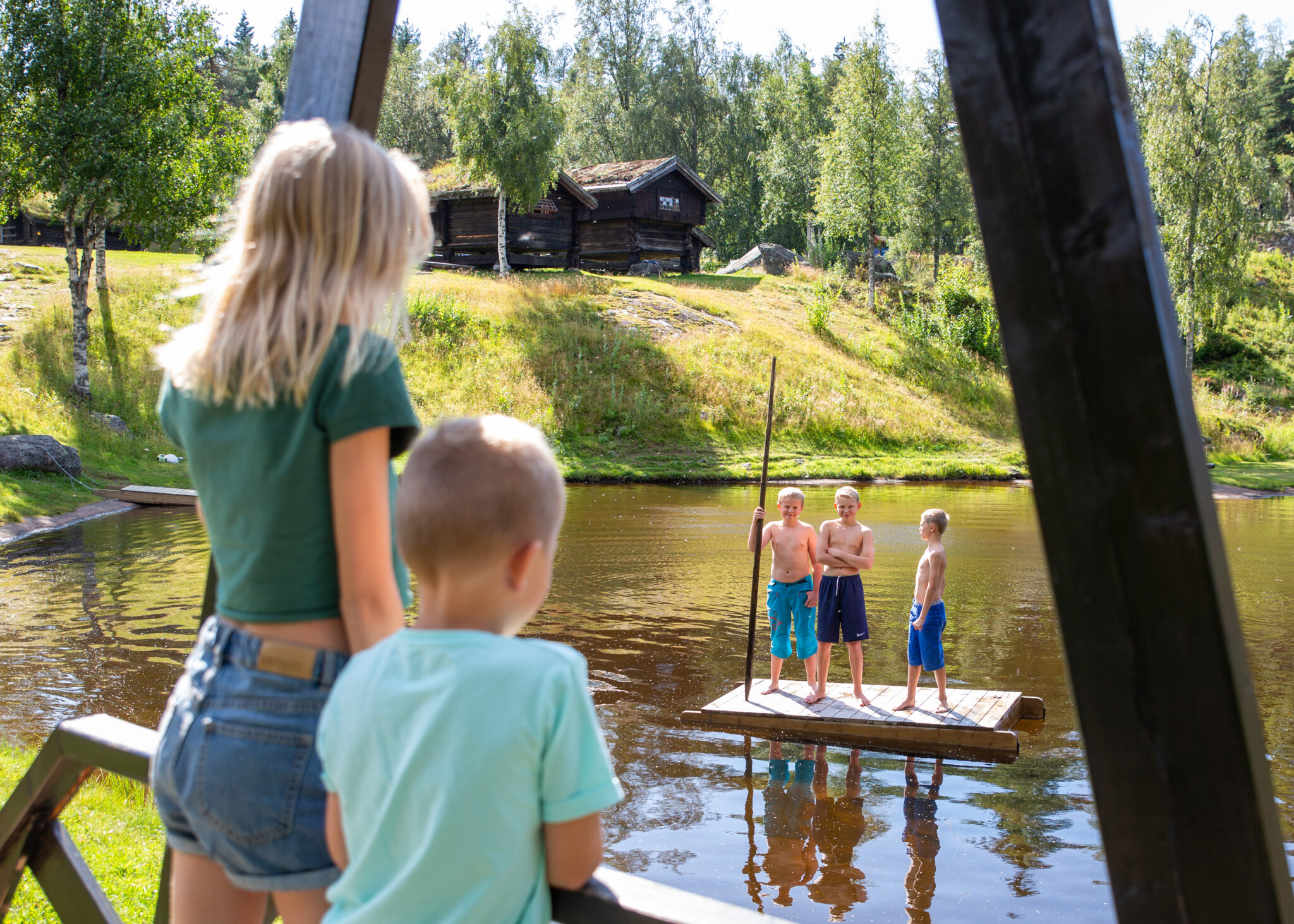 Children stand on a wooden platform in a shallow lake, with shirtless boys in the water and a wooded, cabin-filled backdrop.