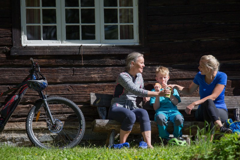 Three people—two women and a boy—sit on a wooden bench outside a log cabin, sharing snacks with a bicycle leaning nearby.