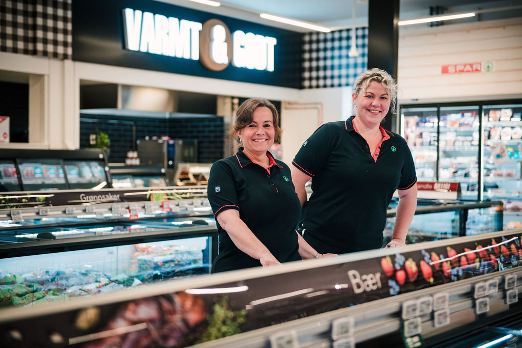 Two store employees in matching polo shirts smile from behind a refrigerated counter in a grocery store.