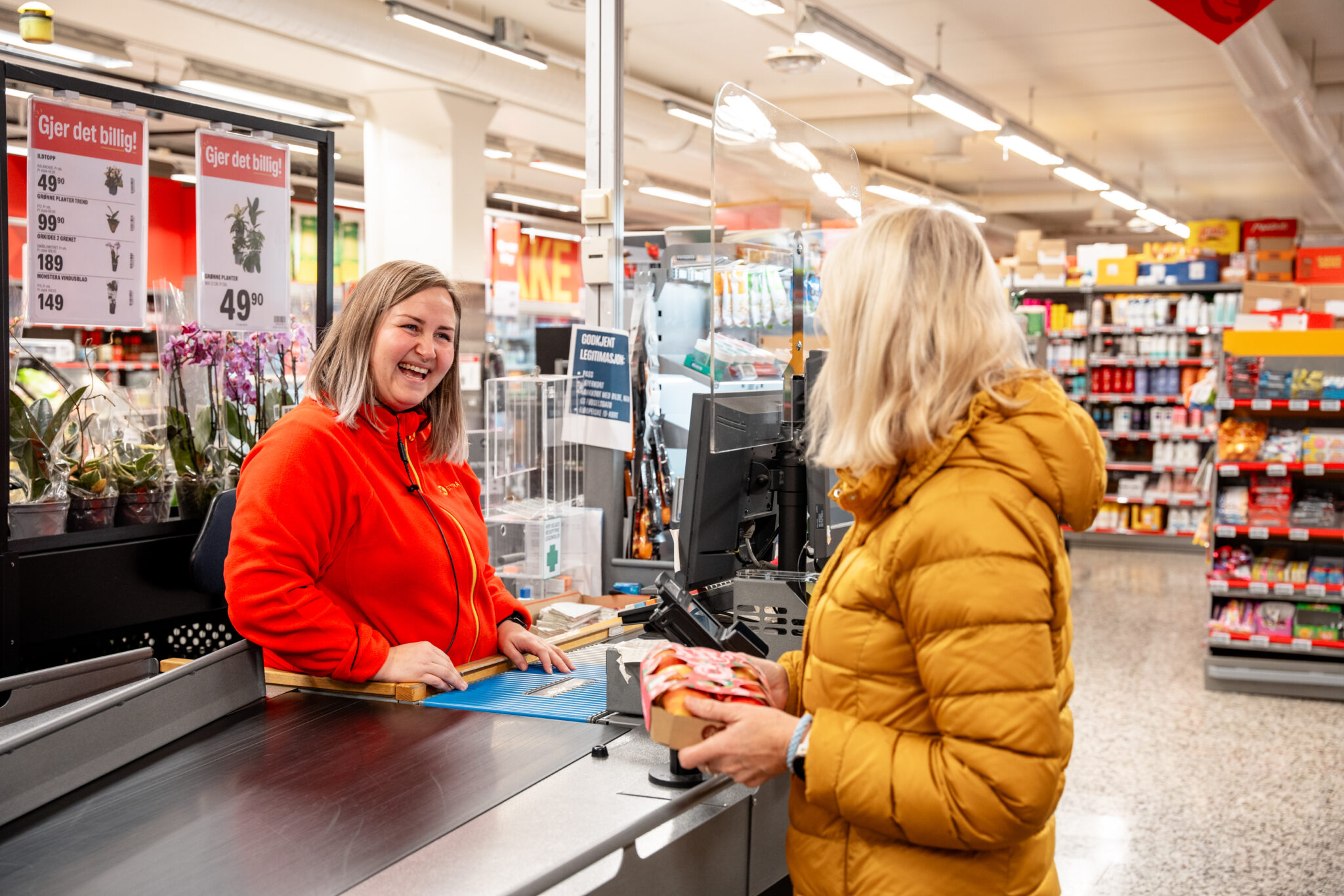 Cashier in red jacket smiling at a checkout while assisting a customer with a product at a supermarket counter.