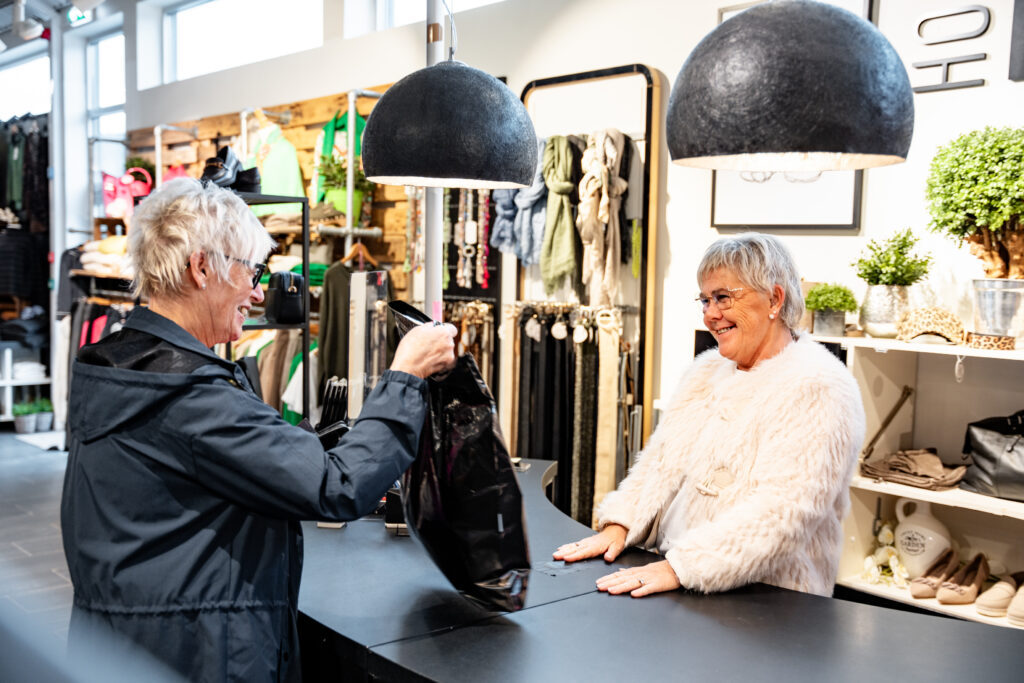 Two elderly women in a clothing store; one in a white fur coat at the counter, the other hands her a black shopping bag.