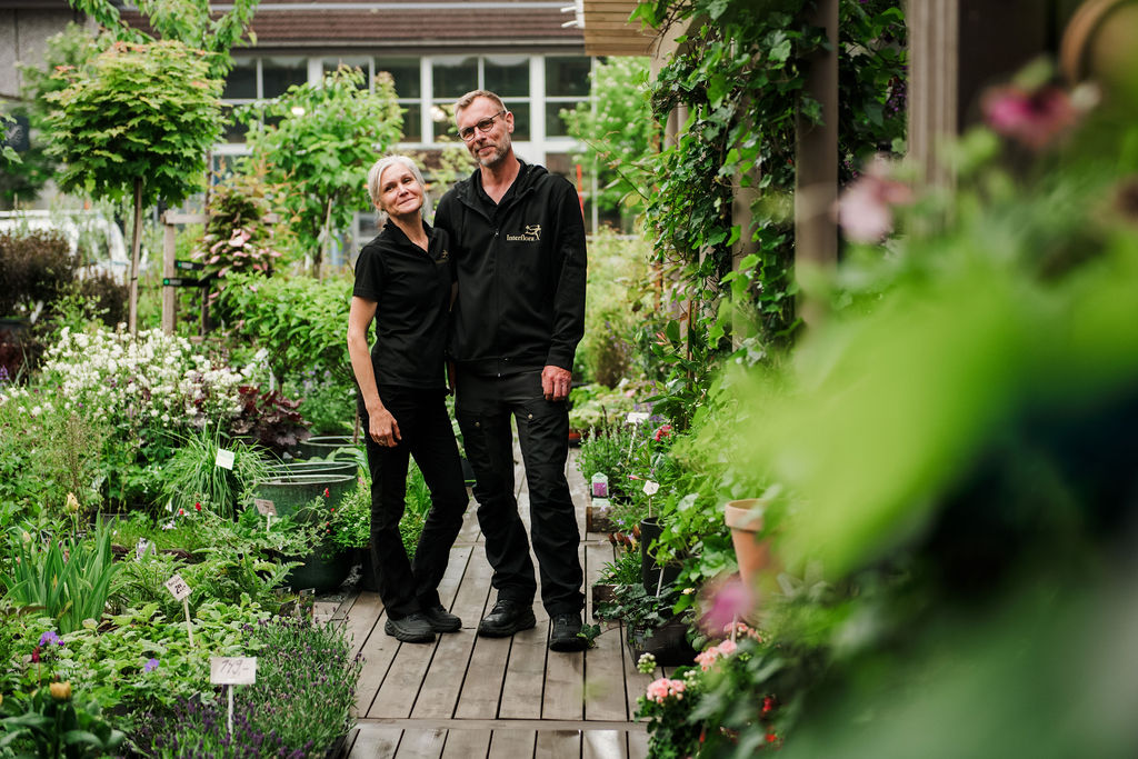 Two gardeners in black pose together on a wooden path amid a lush plant shop/greenhouse.