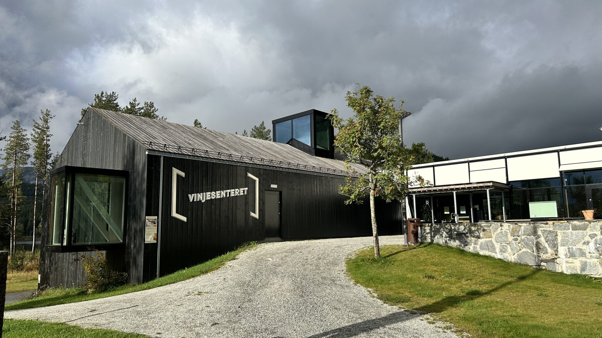 Modern black wooden building with a glass-fronted corner and a sign reading 'VINJESENTERET' beside a gravel path and trees under a cloudy sky.