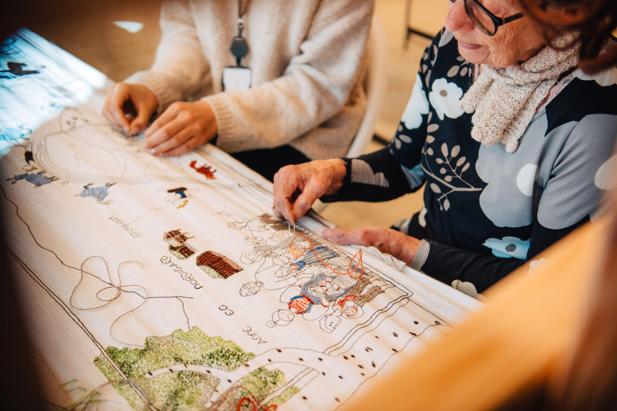 Two seniors sketching on a large illustrated map spread on a table, using pens and markers.