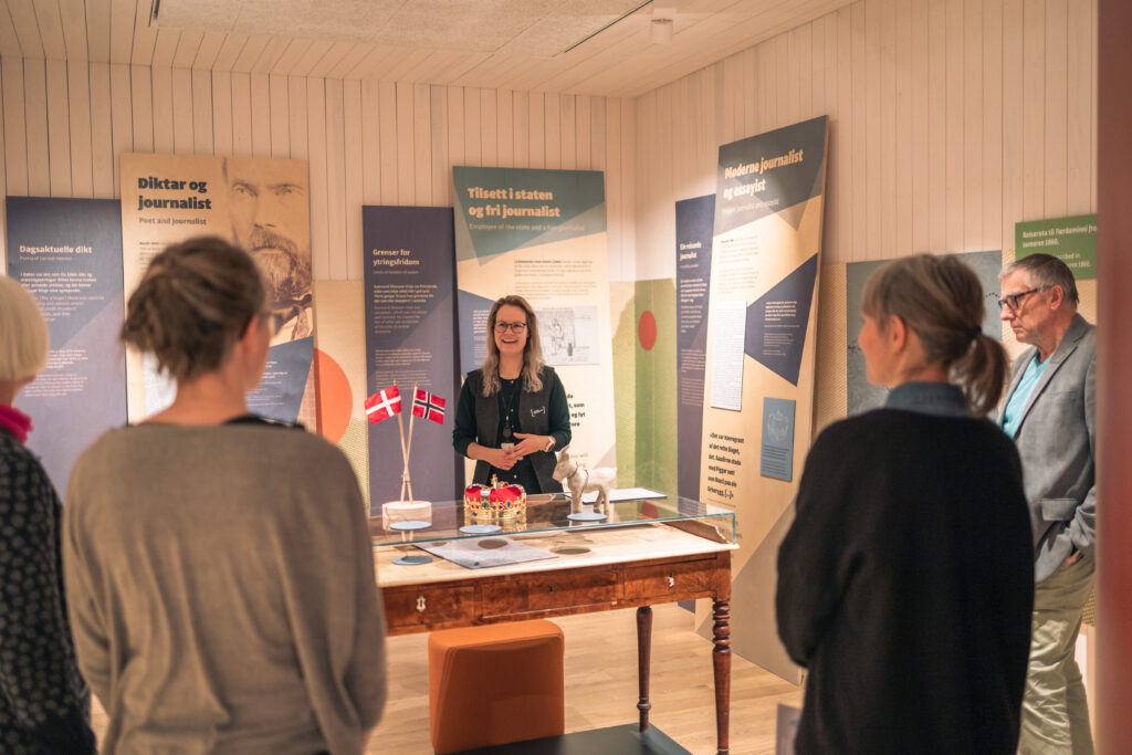Smiling woman presenter standing behind a wooden display case with Danish flags, in a gallery with informational boards on the walls and visitors watching.