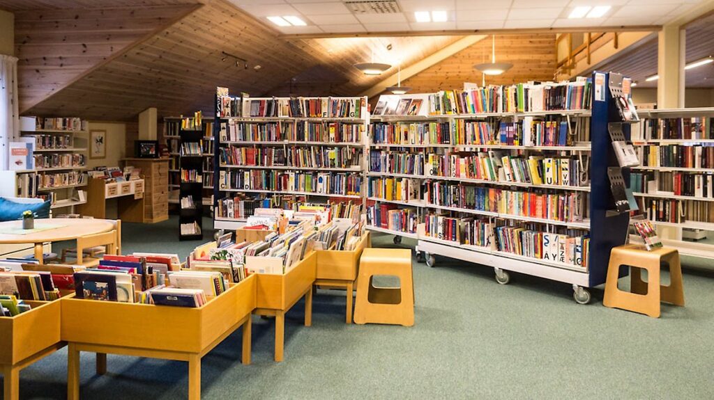 Public library interior with rolling book carts, tall shelves filled with colorful books, and a green carpeted floor.