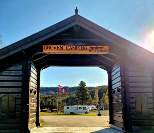 Wooden archway at campsite entrance with a sign reading Groven Camping; blue sky above, Norwegian flag visible.