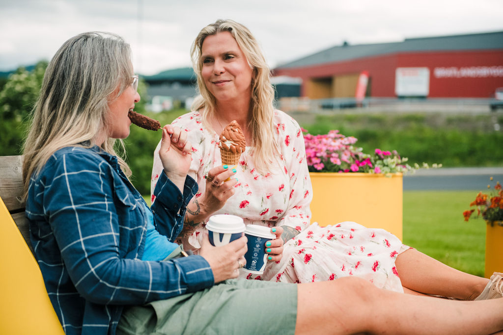 Two women sit on a bench outdoors, sharing ice cream cones and coffee cups, smiling at each other.