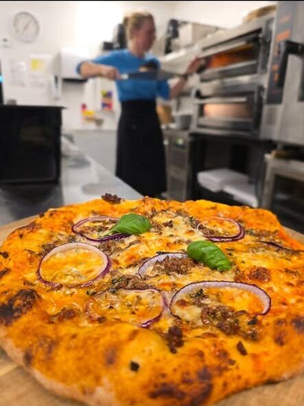 Pizza in the foreground with red onion rings and basil on a wooden counter as a baker removes a tray from a large commercial oven in a busy kitchen.