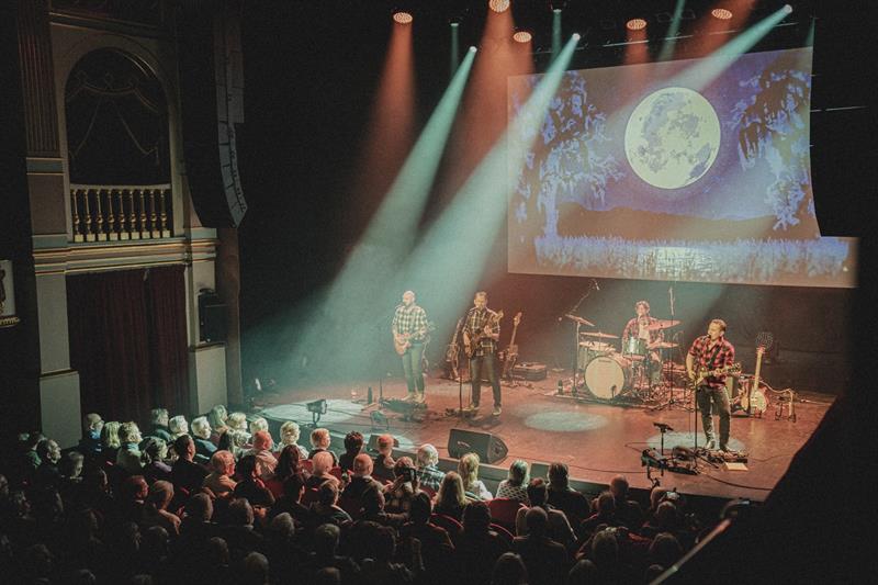 Band performing on stage in a theater with a moon projection on screen and an attentive audience watching from the pit and balconies.