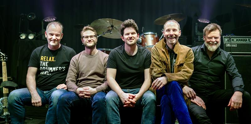 Five men sit on a stage in front of a drum set, posing for a band photo.