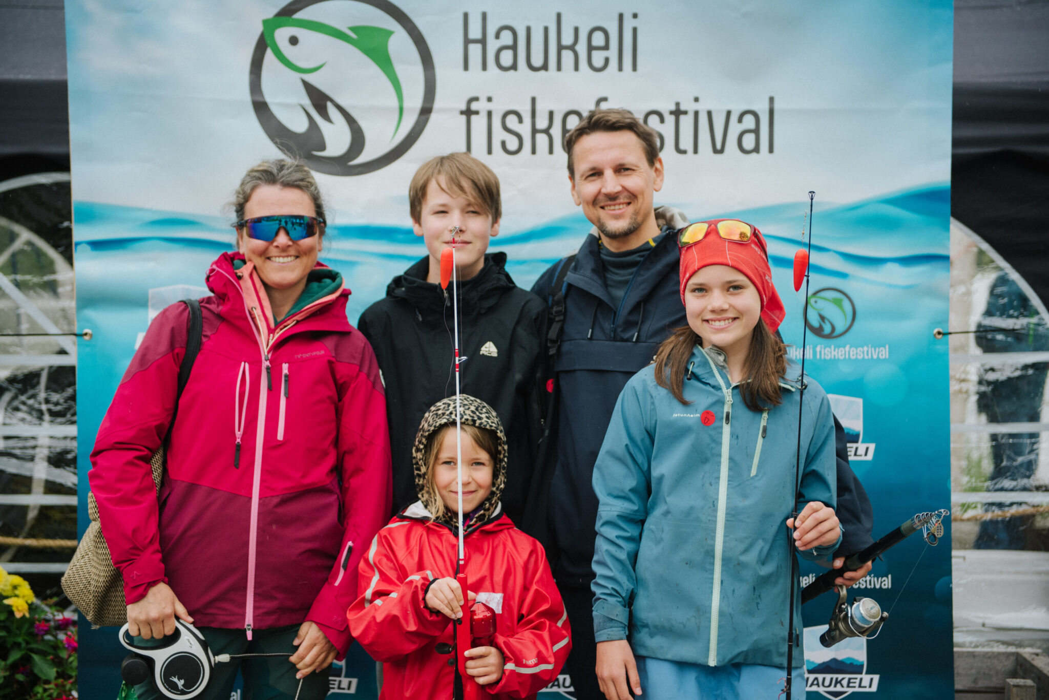Four people in bright outdoor jackets pose and smile in front of a blue festival banner, each holding fishing gear or rods at a fishing festival.
