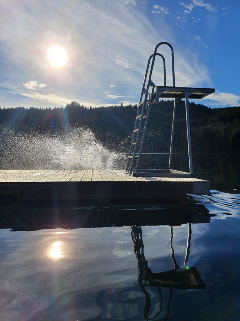 Diving platform on a wooden dock extending over a calm lake with a forested shore and bright sun in a blue sky.
